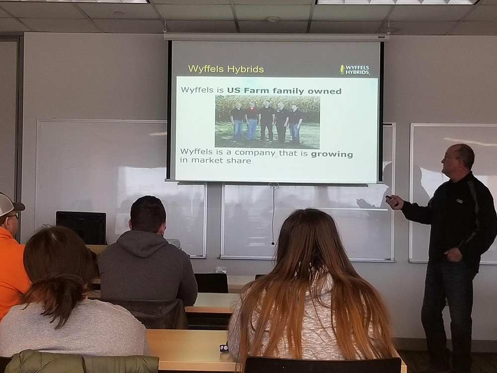 Man standing in front of class pointing to a scren with a PowerPoint slide on it